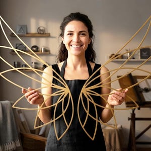 Könnte beinhalten: Eine Frau hält ein Paar goldfarbene, abstrakt geformte Wandkunststücke in Form von Flügeln. Die Kunst zeigt eine Reihe von gebogenen Linien, die in einem symmetrischen Muster angeordnet sind. Der Hintergrund zeigt einen Raum mit Holzregalen und einem Zeichentisch.