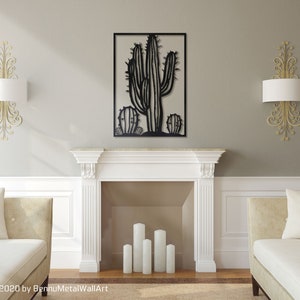 Puede incluir: Arte de pared de metal negro con un cactus grande y dos cactus m&aacute;s peque&ntilde;os a cada lado. El arte se muestra sobre una repisa de chimenea blanca en un ambiente de sala de estar.