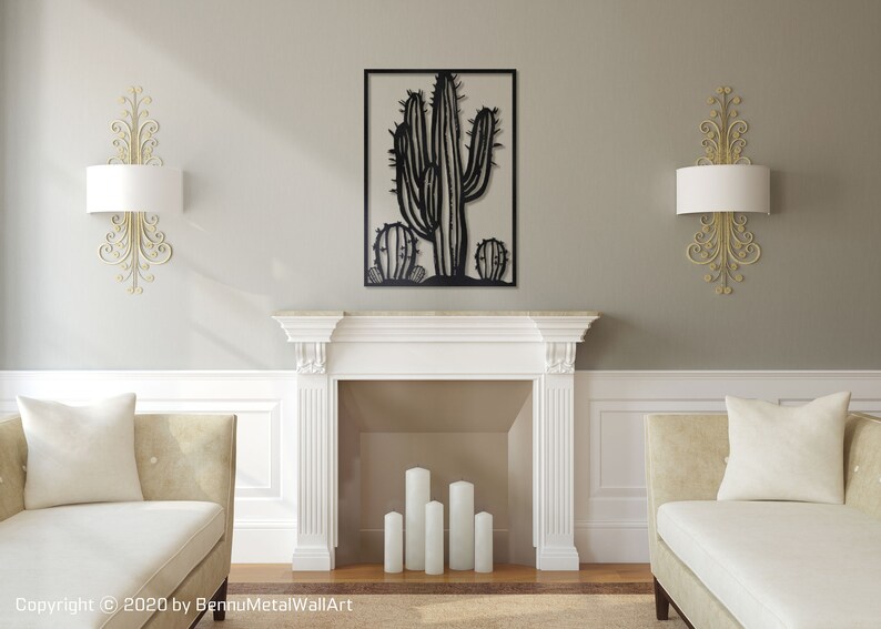 Puede incluir: Arte de pared de metal negro con un cactus grande y dos cactus m&aacute;s peque&ntilde;os a cada lado. El arte se muestra sobre una repisa de chimenea blanca en un ambiente de sala de estar.