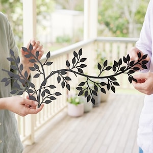 May include: A black metal wall art piece in the shape of a leafy branch. The decorative piece is held by two people. The leaves are cut out of the metal, creating a silhouette effect. The art piece is designed for indoor or outdoor use.