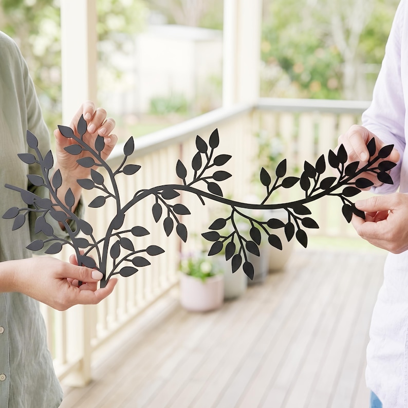 May include: A black metal wall art piece in the shape of a leafy branch. The decorative piece is held by two people. The leaves are cut out of the metal, creating a silhouette effect. The art piece is designed for indoor or outdoor use.