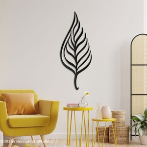 May include: Black metal leaf wall art with a modern design. The leaf is hanging on a white wall in a living room setting with a yellow armchair, side tables, and plants.