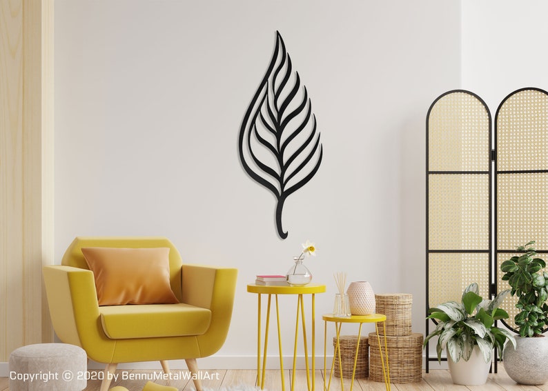 May include: Black metal leaf wall art with a modern design. The leaf is hanging on a white wall in a living room setting with a yellow armchair, side tables, and plants.