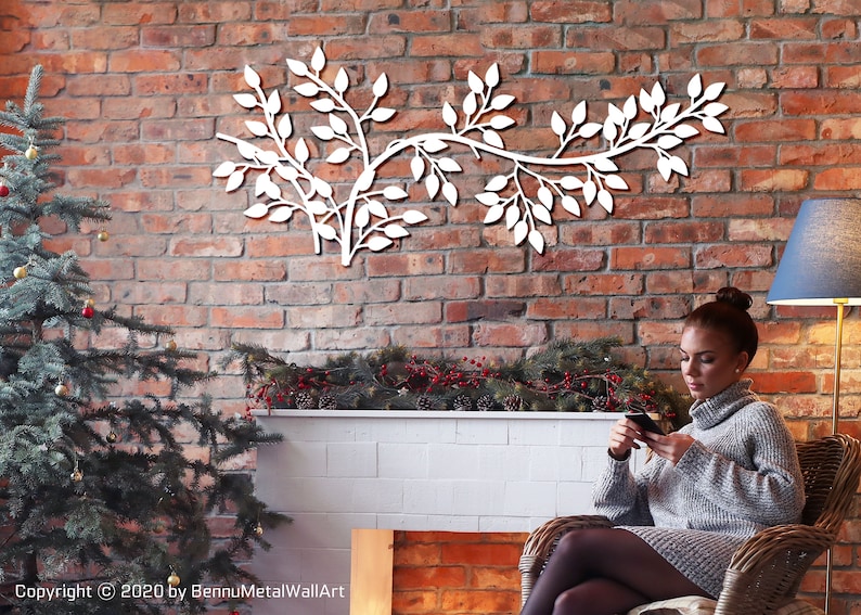 May include: White metal wall art in the shape of a leafy branch, mounted on a red brick wall. The artwork is a decorative element, adding a touch of nature to the interior. The image also includes a Christmas tree and a person.