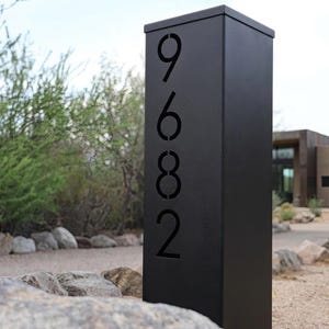 Può includere: Un palo indirizzo nero con i numeri 9682 ritagliati. Il palo è rettangolare e si erge in posizione verticale. I numeri sono in un font moderno. Il palo è ambientato in un ambiente naturale all'aperto con rocce e vegetazione.
