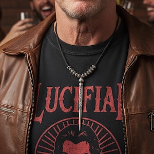 Może przedstawiać: Srebrny naszyjnik z wisiorkiem w kształcie sztyletu. Naszyjnik jest noszony na czarnym t-shircie ze słowem "LUCIFER" w czerwonych literach. Osoba ma na sobie brązową sk&oacute;rzaną kurtkę.