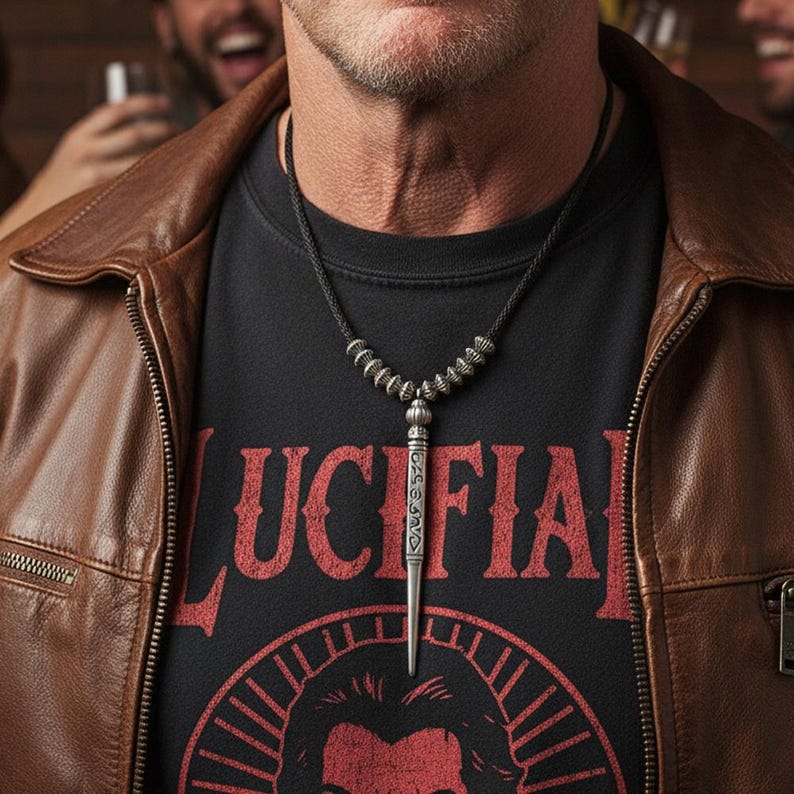 Może przedstawiać: Srebrny naszyjnik z wisiorkiem w kształcie sztyletu. Naszyjnik jest noszony na czarnym t-shircie ze słowem "LUCIFER" w czerwonych literach. Osoba ma na sobie brązową sk&oacute;rzaną kurtkę.