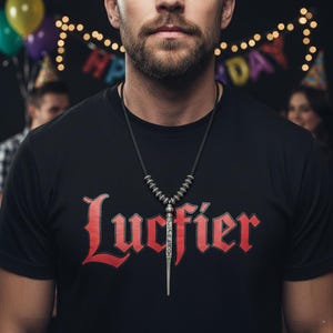 Może przedstawiać: Czarny t-shirt ze słowem "Lucifier" w czerwonym gotyckim kroju pisma. Srebrny naszyjnik z wisiorkiem o długim, spiczastym kształcie wisi na piersi osoby. Dekoracje imprezowe w tle.