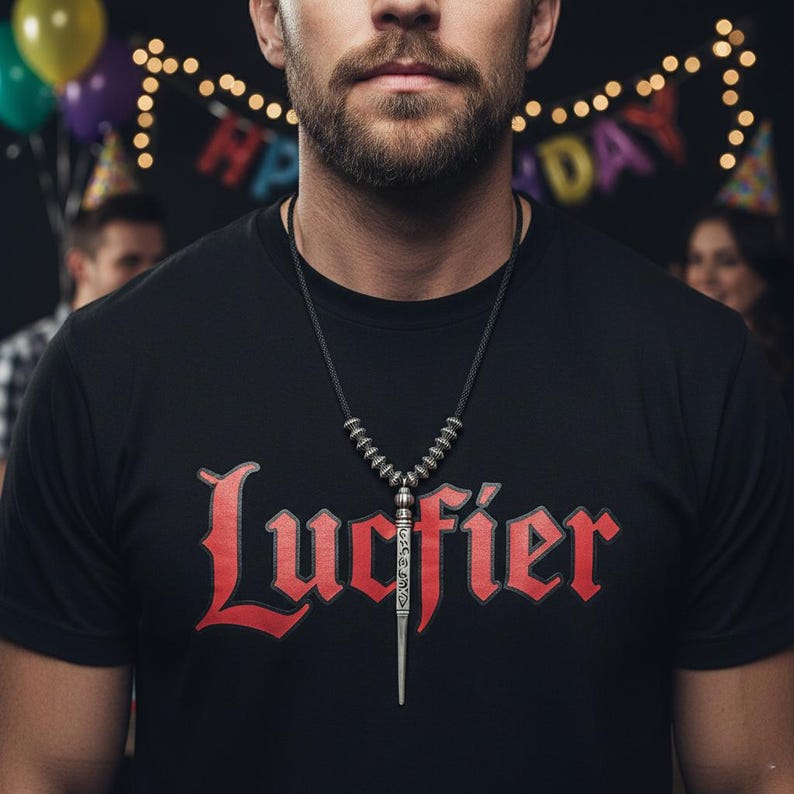 Może przedstawiać: Czarny t-shirt ze słowem "Lucifier" w czerwonym gotyckim kroju pisma. Srebrny naszyjnik z wisiorkiem o długim, spiczastym kształcie wisi na piersi osoby. Dekoracje imprezowe w tle.