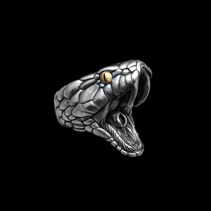 May include: A silver-colored ring shaped like a snake's head with an open mouth. The snake has a detailed scale pattern and a golden-yellow eye. The ring is set against a black background.