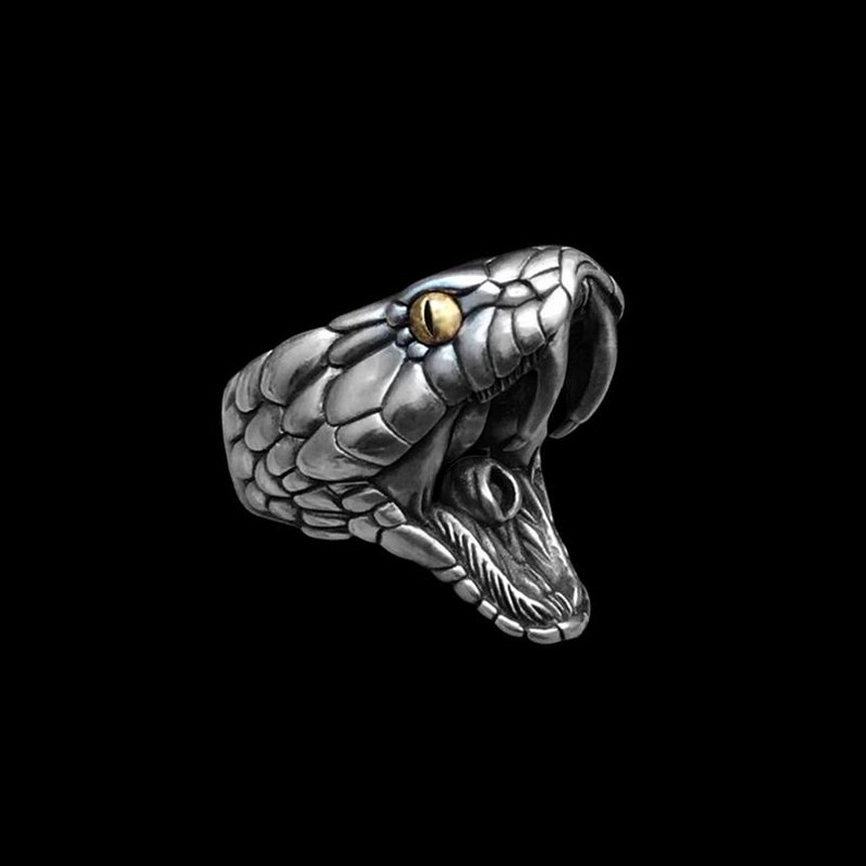 May include: A silver-colored ring shaped like a snake's head with an open mouth. The snake has a detailed scale pattern and a golden-yellow eye. The ring is set against a black background.