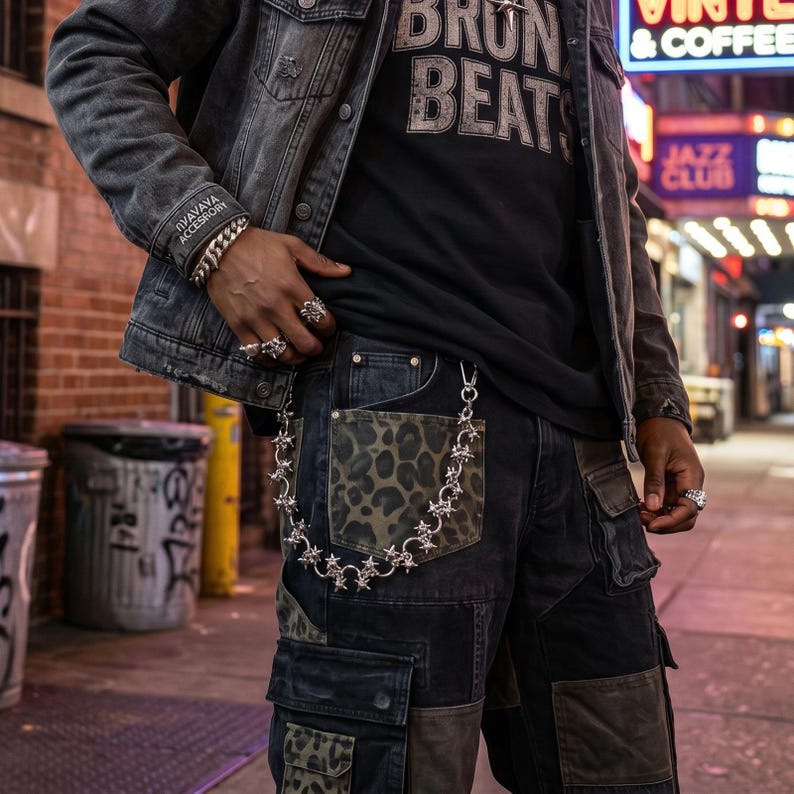 May include: A silver-toned metal chain featuring star-shaped links attached to the pocket of dark-colored cargo pants. The pants include olive green leopard print panels and multiple utility pockets. The chain is secured with a metal clasp.