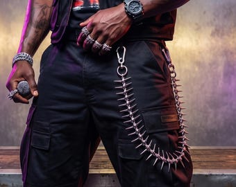 May include: Black cargo pants with silver spiked chain detail. The image also shows a black watch, multiple silver spiked rings, and a silver bracelet. The person is wearing a black vest and a black t-shirt.