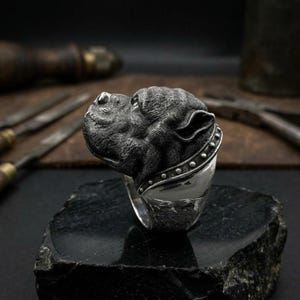 Puede incluir: Un anillo de plata con una detallada escultura de la cabeza de un perro. La cabeza del perro es texturizada y tiene un collar con pequeños tachuelas redondas. El anillo se muestra sobre una superficie negra texturizada.