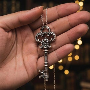 Puede incluir: Collar con colgante de llave plateada. La llave presenta un diseño ornamental y decorativo con un mango detallado y una cadena delicada. La llave se sostiene en una mano, mostrando la intrincada artesanía de la joyería.