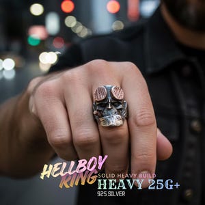 May include: A silver Hellboy ring with a detailed skull design. The ring features copper-colored accents and the text "HELLBOY RING" and "HEAVY 25G+". The ring is made of 925 silver.