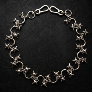 May include: A silver-toned metal chain featuring a series of interconnected circular links, each adorned with a four-pointed star-shaped spike. The chain is finished with two lobster claw clasps at the ends, creating a circular arrangement of hardware.