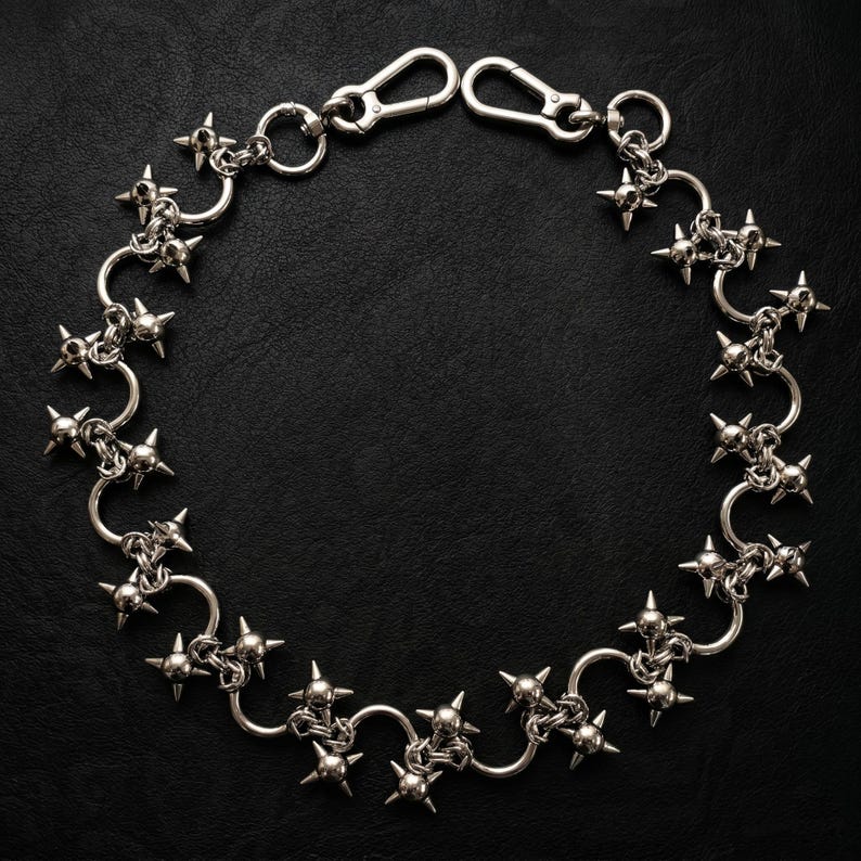 May include: A silver-toned metal chain featuring a series of interconnected circular links, each adorned with a four-pointed star-shaped spike. The chain is finished with two lobster claw clasps at the ends, creating a circular arrangement of hardware.