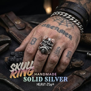 May include: A handmade solid silver skull ring with copper accents. The ring has a detailed design and is heavy, weighing over 25 grams. The image also includes the text "SKULL RING" and "HANDMADE SOLID SILVER HEAVY 25g+".