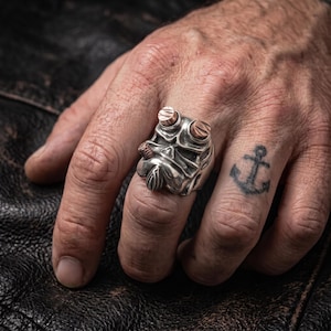 May include: A silver-toned ring featuring a sculpted character face with prominent brow ridges, a central nose detail, and two copper-colored cylindrical accents positioned on the upper portion of the ring band, worn on a finger.