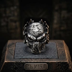 May include: A silver-colored ring featuring a detailed Predator mask design. The ring has a textured surface and is set on a dark, textured base. A small rectangular plaque with engraved symbols is visible below the ring.