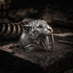Könnte beinhalten: Ein silberner Ring mit einem detaillierten Säbelzahntigerkopf mit offenem Maul und markanten Fangzähnen. Der Ring liegt auf einer dunklen Holzoberfläche, die das aufwendige Design und den metallischen Glanz zeigt.
