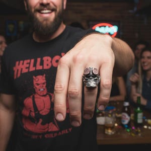 May include: A silver-colored ring with a skull design, featuring red accents on the top. The ring is worn on a hand, with a black t-shirt visible in the background that says "HELLBOY" in red letters.