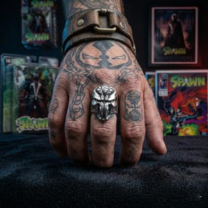 Op de afbeelding: Een zilverkleurige ring met een gedetailleerd schedelontwerp wordt getoond op een getatoeëerde hand. De ring is het middelpunt, met strips en actiefiguren met het personage Spawn op de achtergrond. Een bruine leren polsband is ook zichtbaar.