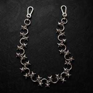 May include: A silver-toned metal chain featuring a series of interconnected circular links, each adorned with multiple sharp, protruding spikes. The chain is finished with a lobster claw clasp at each end, designed for secure attachment to garments.