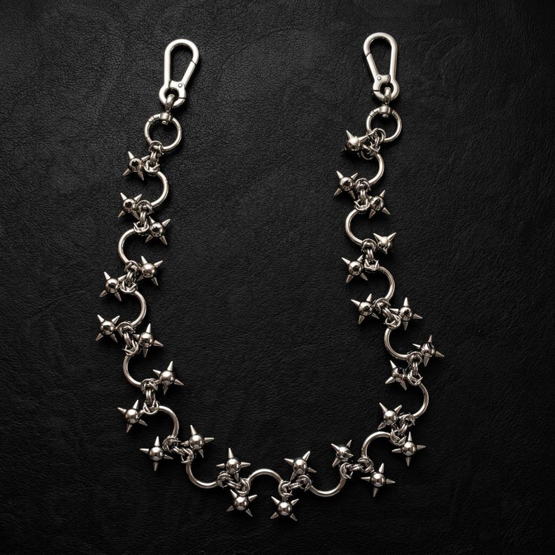 May include: A silver-toned metal chain featuring a series of interconnected circular links, each adorned with multiple sharp, protruding spikes. The chain is finished with a lobster claw clasp at each end, designed for secure attachment to garments.