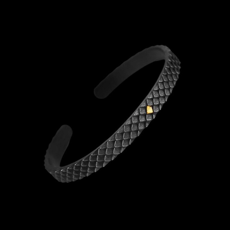 Puede incluir: Una pulsera de metal negro con un dise&ntilde;o de piel de serpiente texturizado y un peque&ntilde;o acento dorado.