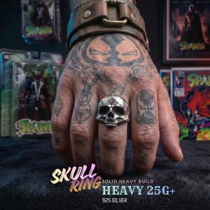 May include: A silver skull ring with a geometric design is shown on a hand with tattoos. The ring is labelled "SKULL RING" and "HEAVY 25G+" with "925 SILVER" below. The background includes comic books and action figures.