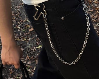 May include: A silver-colored chain wallet accessory attached to black pants. The chain is connected to a decorative cross-shaped clasp at the belt loop. The person is wearing a white shirt and holding a black bag.