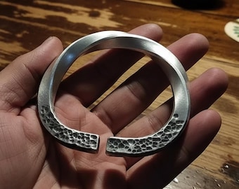 May include: A silver-colored, open-ended bracelet with a textured, hammered design on the ends. The bracelet is held in a hand, showcasing its smooth, curved form and the detailed surface texture.