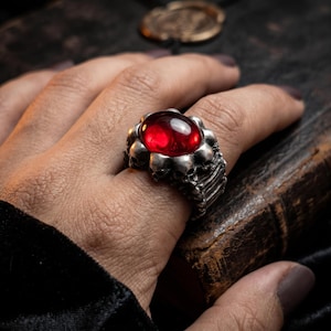 May include: A silver ring featuring a large, oval, red gemstone surrounded by small skull designs. The ring is worn on a hand, with the details of the ring's design clearly visible. The ring has a gothic or fantasy theme.