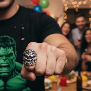 Puede incluir: Un anillo plateado con el rostro esculpido de Hulk. El anillo se lleva en un puño cerrado. El fondo muestra a una persona con una camiseta negra con un gráfico de Hulk y una escena de fiesta.