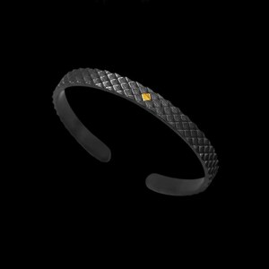 Puede incluir: Una pulsera de metal negro con un dise&ntilde;o texturizado escamoso y un peque&ntilde;o acento cuadrado dorado.