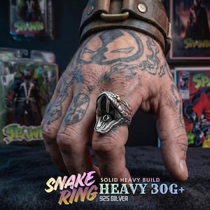 May include: A silver snake ring with an open mouth design, featuring red and gold accents. The ring is held up by a hand with tattoos. The text "SNAKE RING" and "HEAVY 30G+" is visible.