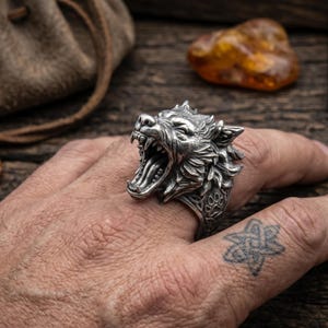 May include: A silver ring with a detailed wolf head design, showing the wolf with its mouth open. The ring has intricate detailing on the wolf's fur and a paw print design on the band. The ring is shown on a hand.