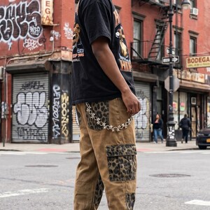 May include: A black short-sleeved t-shirt with a graphic print and tan cargo pants featuring leopard print panels on the pockets and sides. A silver-toned metal chain with star-shaped links is attached to the waistband of the pants.