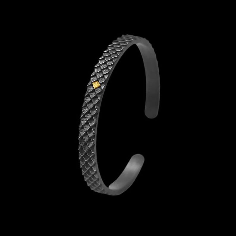 Puede incluir: Un brazalete de metal negro con un dise&ntilde;o texturizado de piel de serpiente y un peque&ntilde;o acento cuadrado dorado.