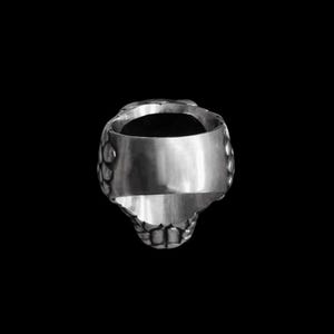 May include: A silver-colored ring with a skull design. The ring features a detailed skull shape with textured patterns on the sides and a smooth band. The ring is set against a black background.
