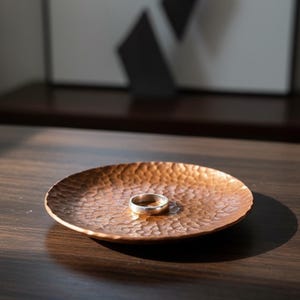 May include: A hammered copper ring dish with a silver ring resting in the center. The dish has a textured surface and is round. The dish is sitting on a dark wooden surface. The background includes a dark wooden shelf and a piece of art.