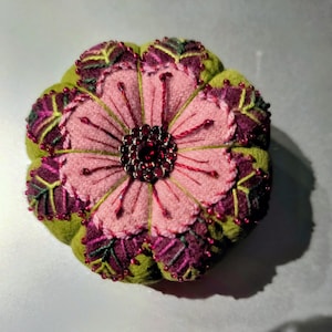 May include: A round, multi-layered fabric flower ornament featuring a central cluster of dark red crystals. The petals are crafted from light pink, deep purple, and lime green felt, accented with intricate embroidery and small red beads.