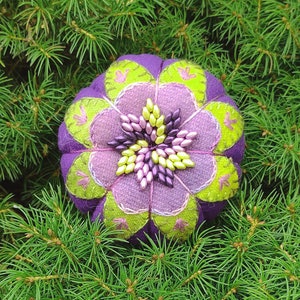 Puede incluir: Un cojín de alfileres de tela morado y verde con un centro en forma de estrella hecho de cuentas rosas y verdes.