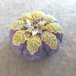 Pode incluir: Um almofadinha para alfinetes roxo com um design de flor feito de tecido verde e rosa e contas brancas, azuis e douradas.