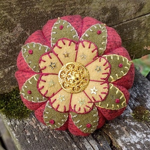 Pode incluir: Um almofadinha para alfinetes vermelho, verde e dourado com um design de estrela e um botão dourado no centro.