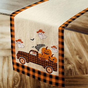 May include: A beige table runner with a black and orange plaid border. The runner features a vintage truck carrying a stack of pumpkins and ghosts with hats. The text "Boo!" is printed on the runner.