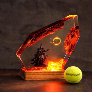 May include: A resin sculpture featuring a dark figure and a golden ring within a fiery, red and orange landscape. The sculpture is set on a wooden base and illuminated with warm light. A yellow tennis ball with the text "Sonbioukl" is in the foreground.