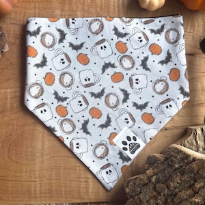 May include: A triangular pet bandana featuring a white background with a repeating pattern of orange pumpkins, black bats, and white ghost-shaped mugs. A small white fabric tag with a black paw print logo is attached to the lower right corner.